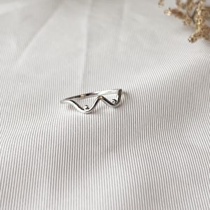 May include: A silver ring with a wavy design. The ring features three curved sections with small, round accents. The ring is set against a white, striped background.