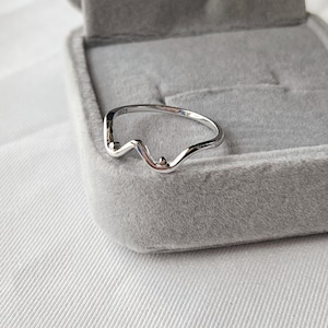 May include: A silver-colored ring with a unique, wavy design. The ring features a small, raised detail and is set against a gray velvet jewelry box. The ring is a minimalist piece of jewelry.