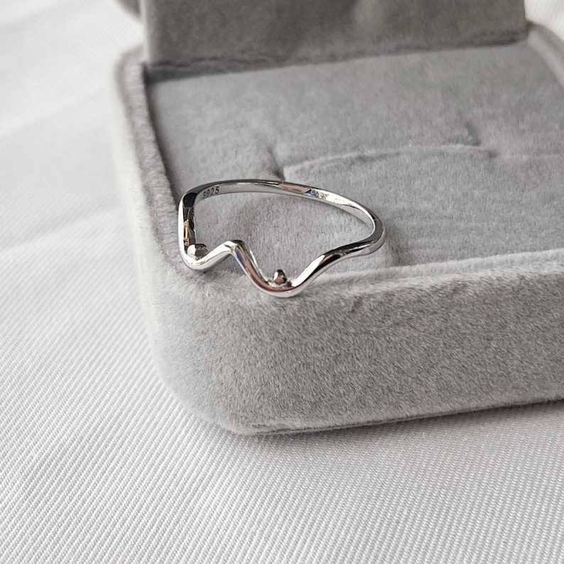 May include: A silver-colored ring with a unique, wavy design. The ring features a small, raised detail and is set against a gray velvet jewelry box. The ring is a minimalist piece of jewelry.