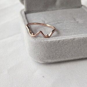 May include: Rose gold ring with a wavy design and two small, round accents. The ring is set against a gray velvet jewelry box, with a white fabric background. The ring's unique shape adds a touch of elegance.