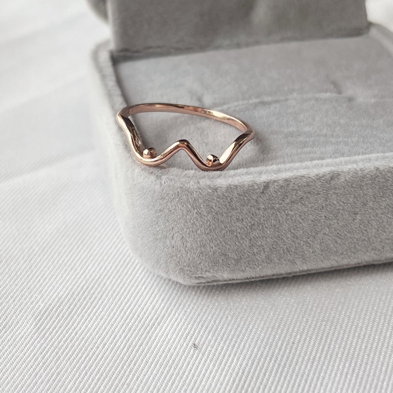May include: Rose gold ring with a wavy design and two small, round accents. The ring is set against a gray velvet jewelry box, with a white fabric background. The ring's unique shape adds a touch of elegance.