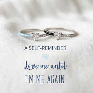 Puede incluir: Dos anillos de plata con piedras centrales en forma de corazón y pequeñas piedras de acento. Un anillo tiene una banda de esmalte azul claro, el otro una banda de esmalte negro. El texto dice "A SELF-REMINDER" y "Love me until I'M ME AGAIN."
