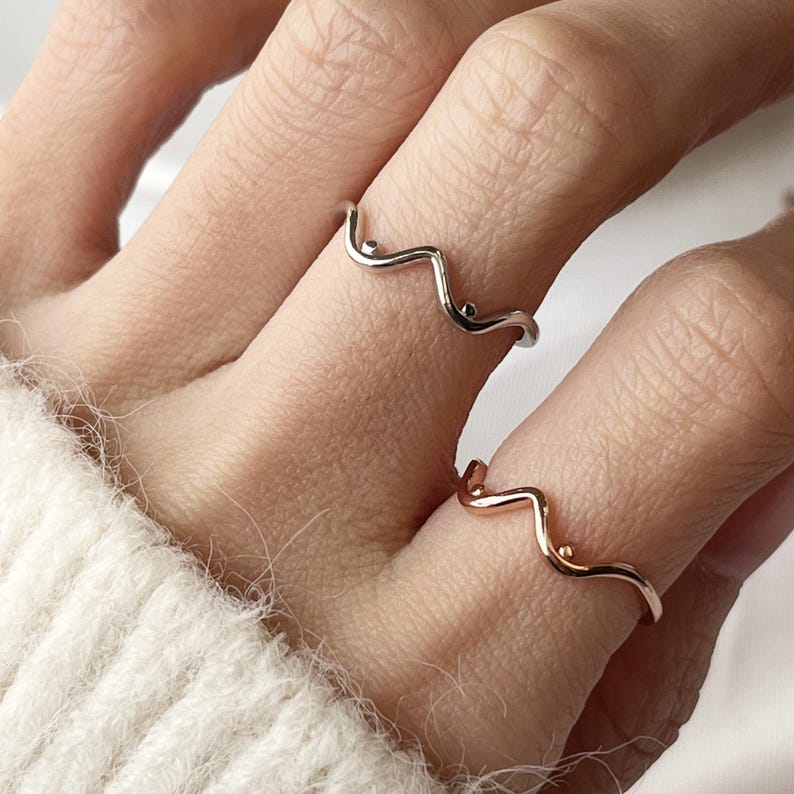 May include: Two delicate wave-shaped rings. One ring is silver-toned, and the other is rose gold. Each ring features a minimalist design with small, round accents. The rings are worn on fingers, showcasing their elegant and simple style.