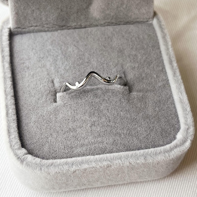 May include: A silver-colored ring with a wavy design, presented in a gray velvet ring box. The ring features a delicate, flowing shape with small, round accents. The jewelry is displayed against a soft, neutral background.