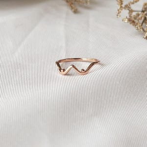 May include: A rose gold ring with a unique wave design. The ring features a delicate, minimalist style with a curved shape and small, round accents. The jewelry is set against a white background, highlighting its elegant form.
