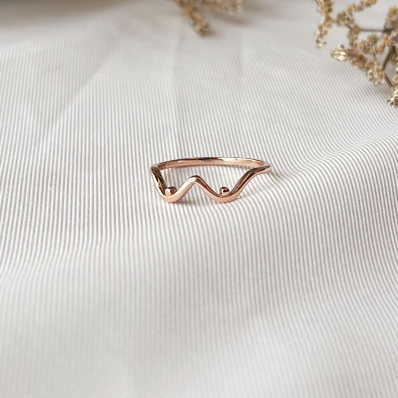 May include: A rose gold ring with a unique wave design. The ring features a delicate, minimalist style with a curved shape and small, round accents. The jewelry is set against a white background, highlighting its elegant form.