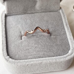 May include: A rose gold ring with a wave design, set in a gray velvet ring box. The ring has a delicate, curved shape with small, round accents. The box is open, showcasing the ring's elegant simplicity.