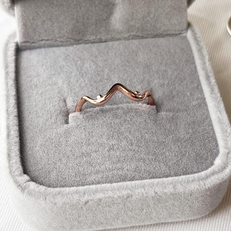 May include: A rose gold ring with a wave design, set in a gray velvet ring box. The ring has a delicate, curved shape with small, round accents. The box is open, showcasing the ring's elegant simplicity.