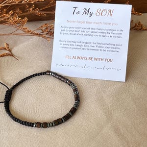 Könnte beinhalten: Ein schwarzes geflochtenes Armband mit Holz- und silberfarbenen Perlen wird neben einer Karte gezeigt. Die Karte trägt die Aufschrift "To My Son" mit einer inspirierenden Botschaft. Das Armband ist verstellbar.