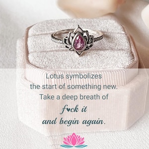 Può includere: Un anello d'argento con un design a fiore di loto, con una gemma rosa al centro. L'anello è esposto in un portagioie rosa chiaro. Il testo sull'immagine recita: "Il loto simboleggia l'inizio di qualcosa di nuovo. Fai un respiro profondo e ricomincia."