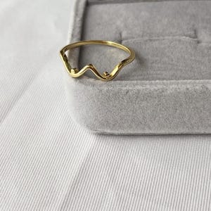 May include: A gold-colored ring with a wavy design. The ring has a delicate, minimalist style, featuring a series of curves and small, round accents. The ring is set against a gray velvet jewelry box and a white fabric background.