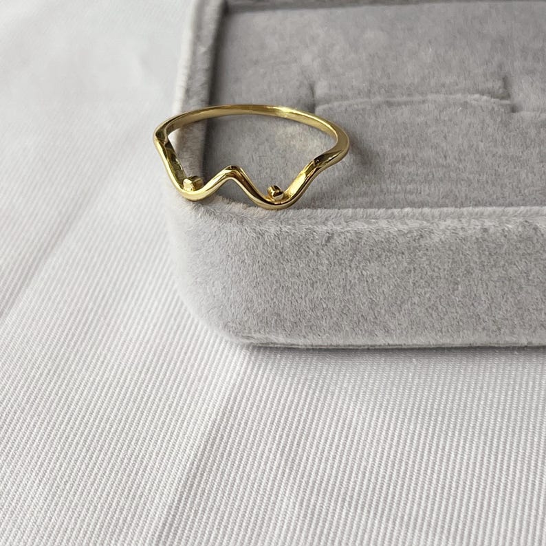 May include: A gold-colored ring with a wavy design. The ring has a delicate, minimalist style, featuring a series of curves and small, round accents. The ring is set against a gray velvet jewelry box and a white fabric background.
