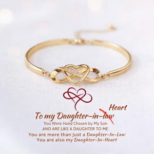 Puede incluir: Un brazalete dorado con un colgante en forma de corazón entrelazado con un símbolo de infinito, adornado con pequeñas piedras transparentes. El brazalete se muestra sobre una superficie blanca texturizada. El texto "To my Daughter-in-law" está presente.