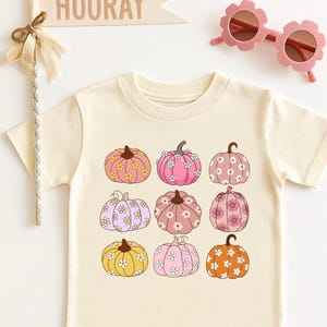 May include: A cream-colored t-shirt featuring a graphic print of nine pumpkins in various shades of pink, orange, and yellow, each decorated with small white flower patterns. Above the shirt is a beige pennant flag with the text HOORAY.