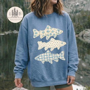 May include: A blue crewneck sweatshirt featuring three stacked fish graphics. The top two fish have a cream background with a blue floral pattern, while the bottom fish has a light blue and white gingham check pattern. The sweatshirt has long sleeves.