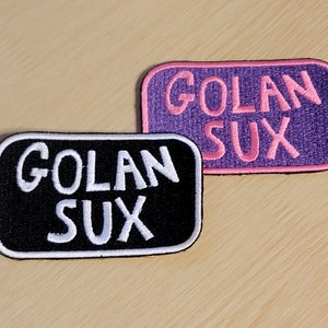 Puede incluir: Dos parches bordados con el texto "Golan Sux" en letras blancas. El parche superior es morado con letras rosas y el parche inferior es negro con letras blancas.