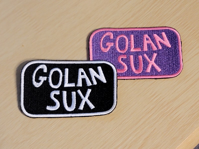 Puede incluir: Dos parches bordados con el texto "Golan Sux" en letras blancas. El parche superior es morado con letras rosas y el parche inferior es negro con letras blancas.