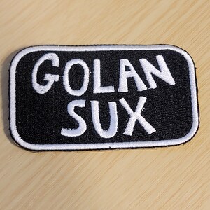 May include: Black and white embroidered patch with the text "Golan Sux" in white lettering.