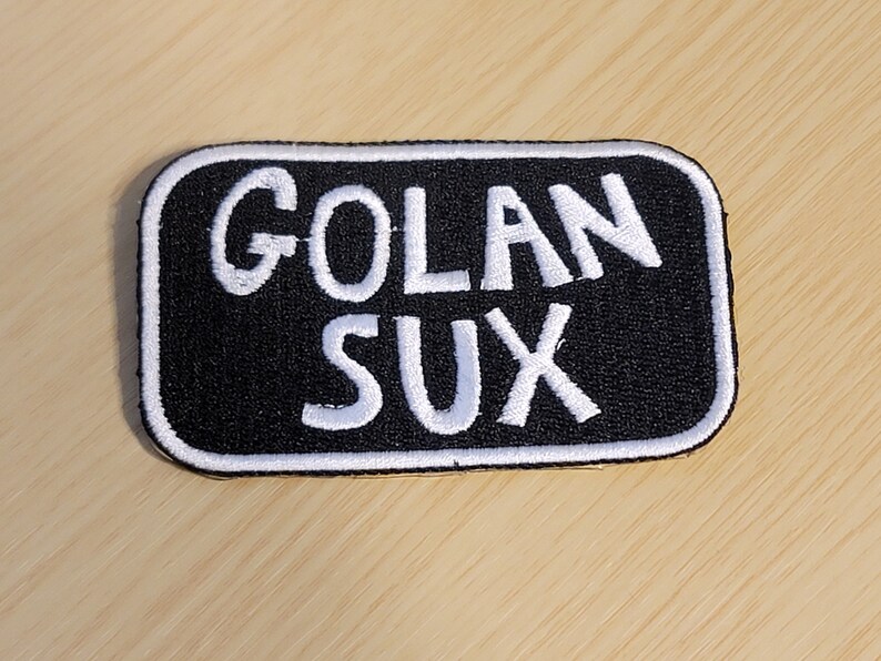 Puede incluir: Parche bordado en blanco y negro con el texto "Golan Sux" en letras blancas.