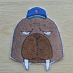 Puede incluir: Parche bordado con un dibujo de una morsa con una gorra de béisbol azul. La morsa tiene un cuerpo marrón, un gran hocico marrón y colmillos blancos. El parche tiene un borde blanco.
