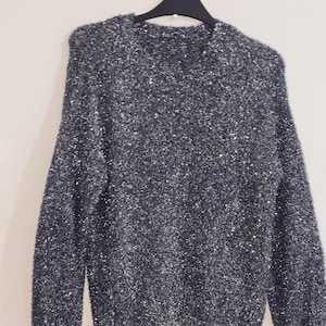 May include: A dark grey sweater with a fuzzy texture and sparkling silver threads. The long-sleeved garment is hanging on a black hanger against a plain white background. The sweater has a crew neck and ribbed cuffs.