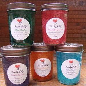 May include: Five glass jars filled with colorful jelly, each with a white label and a red heart. The labels read "Smelly Jelly!" and list the scent of each jar: Mint Medley, Black Cherry Cream, Vanilla Lavender Chamomile, Orange Pakse Tea, and Blue Hawaii.