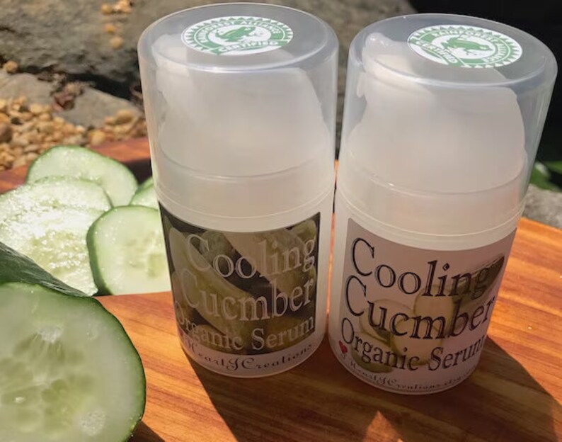 May include: Two clear plastic bottles of Cooling Cucumber Organic Serum with green labels. The labels have a green frog logo and the text "Cooling Cucumber Organic Serum". The bottles are sitting on a wooden surface with sliced cucumbers.