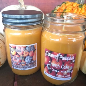 May include: Two glass jars with lids filled with orange soy candles. The labels on the jars read "Caramel Pumpkin Crunch Cake Soy Candle with Hemp Cotton Wick". The jars are surrounded by small white and orange pumpkins.