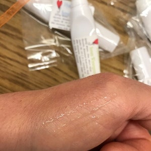 May include: A clear, viscous liquid is applied to a person's hand. The liquid is being applied from a white tube with a black cap. The tube has a label with the text "Lemon & Ginger" on it.