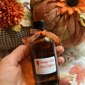 May include: A brown glass bottle labeled "Orange Clove Reed Diffuser" with a black cap and orange ribbon. The bottle is held in a hand, with a fall-themed background featuring decorative pumpkins and flowers.