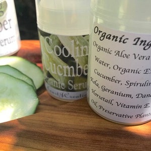 May include: Two clear glass bottles with white labels. The labels read "Cooling Cucumber Organic Serum" and "Organic Ingredients: Organic Aloe Vera Gel, Spring Water, Organic Extracts of Cucumber, Spirulina, Gotu Kola, Geranium, Dandelion, Horsetail, Vitamin E, Jojoba Oil, Preservative Plant". The bottles are sitting on a wooden surface with sliced cucumbers.