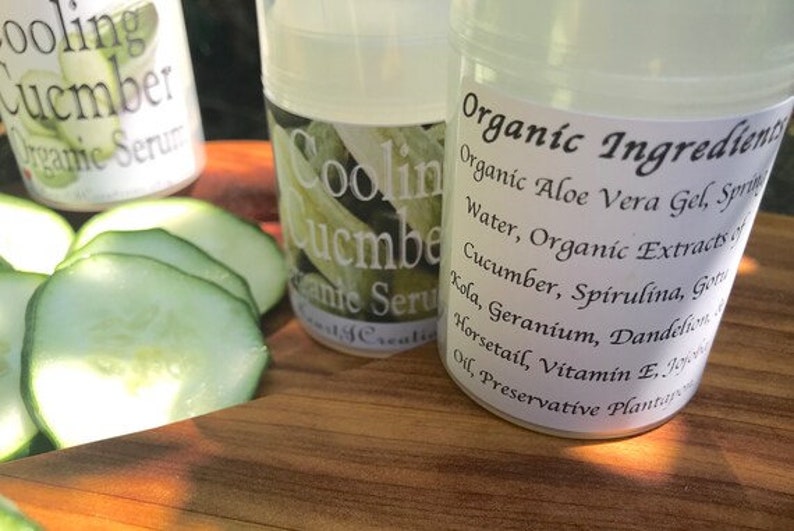 May include: Two clear glass bottles with white labels. The labels read "Cooling Cucumber Organic Serum" and "Organic Ingredients: Organic Aloe Vera Gel, Spring Water, Organic Extracts of Cucumber, Spirulina, Gotu Kola, Geranium, Dandelion, Horsetail, Vitamin E, Jojoba Oil, Preservative Plant". The bottles are sitting on a wooden surface with sliced cucumbers.