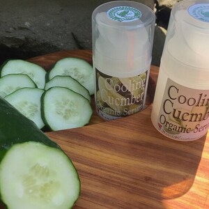May include: Two clear plastic bottles of cooling cucumber organic serum on a wooden surface with sliced cucumbers. The bottles have green labels with the text "Cooling Cucumber Organic Serum".