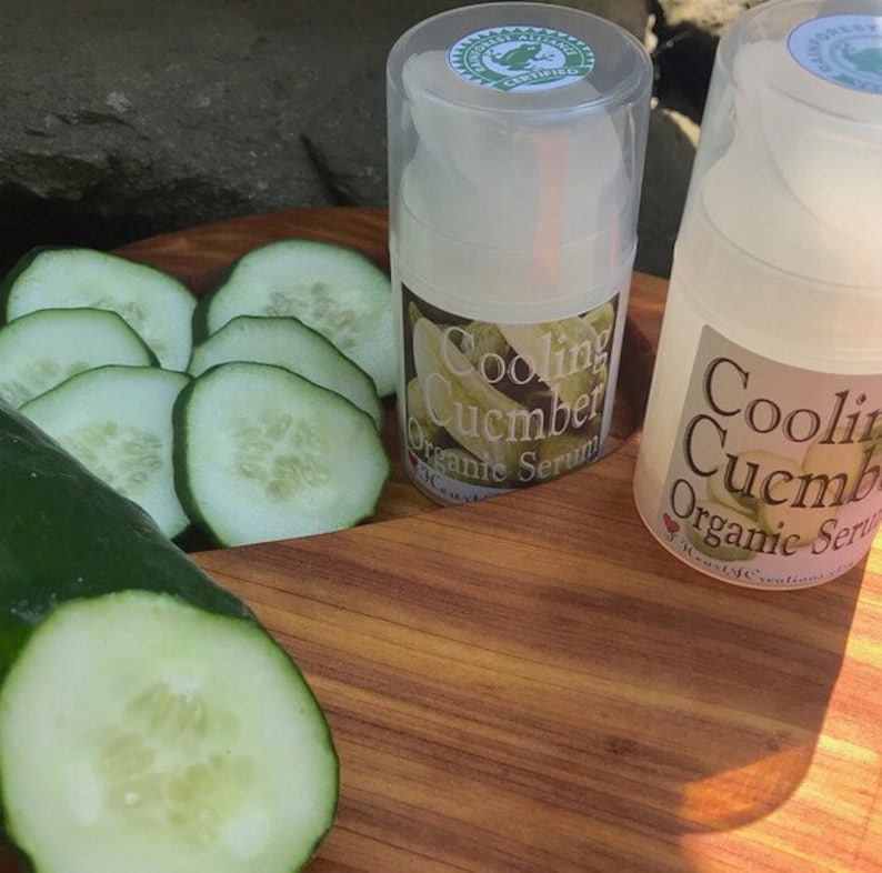 May include: Two clear plastic bottles of cooling cucumber organic serum on a wooden surface with sliced cucumbers. The bottles have green labels with the text "Cooling Cucumber Organic Serum".