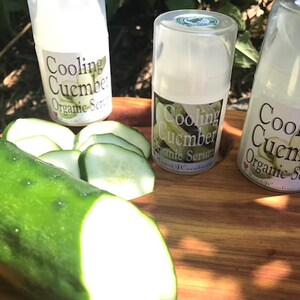 May include: Three small bottles of cooling cucumber organic serum on a wooden surface with a whole cucumber and sliced cucumbers.