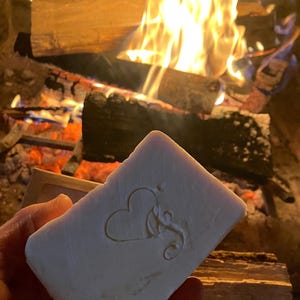 Puede incluir: Una barra de jab&oacute;n blanca con un coraz&oacute;n y un dise&ntilde;o floral en relieve en la parte delantera. El jab&oacute;n se est&aacute; sosteniendo frente a una chimenea con fuego.