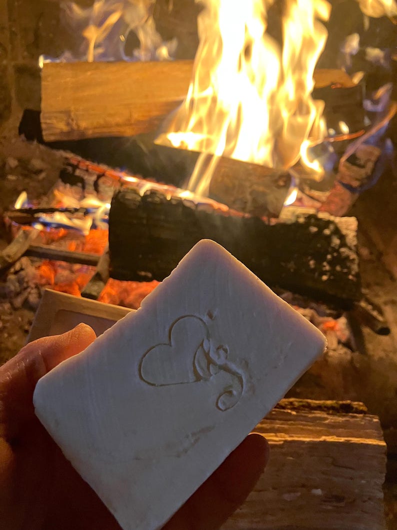 Puede incluir: Una barra de jab&oacute;n blanca con un coraz&oacute;n y un dise&ntilde;o floral en relieve en la parte delantera. El jab&oacute;n se est&aacute; sosteniendo frente a una chimenea con fuego.