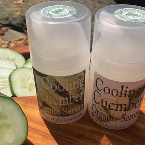 May include: Two clear plastic bottles of Cooling Cucumber Organic Serum with green labels. The labels have a green frog logo and the text "Cooling Cucumber Organic Serum". The bottles are sitting on a wooden surface with sliced cucumbers.