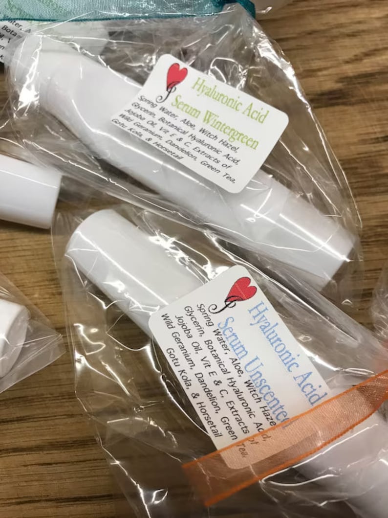 May include: Two clear plastic bags containing white tubes with labels that read "Hyaluronic Acid Serum Wintergreen" and "Hyaluronic Acid Serum Unscented". The labels list ingredients including spring water, aloe, witch hazel, glycerin, botanical hyaluronic acid, jojoba oil, vitamin E and C extracts of wild geranium, dandelion, green tea, gotu kola, and horsetail.