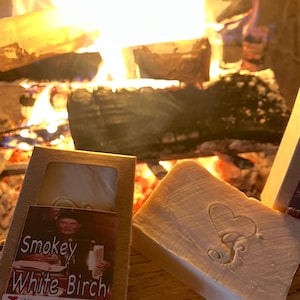 Puede incluir: Dos barras de jab&oacute;n hechas a mano, una con un dise&ntilde;o de coraz&oacute;n y la otra con una etiqueta que dice "Smokey White Birch" y "Heartfelt Creations". El jab&oacute;n se muestra frente a una chimenea con un fuego crepitante.