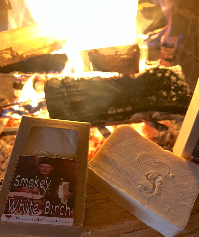 Puede incluir: Dos barras de jab&oacute;n hechas a mano, una con un dise&ntilde;o de coraz&oacute;n y la otra con una etiqueta que dice "Smokey White Birch" y "Heartfelt Creations". El jab&oacute;n se muestra frente a una chimenea con un fuego crepitante.