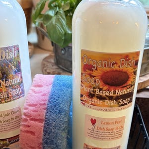 May include: Two clear plastic bottles of Organic Dish Soap, each 16 ounces, featuring labels with sunflower imagery and text. One label reads Lemon Peel Dish Soap. A dual-sided pink and blue sponge sits between the bottles in a black wire basket.