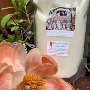 May include: A clear plastic bag filled with white Goat Milk Conditioner. The label reads "Goat Milk Conditioner" with "Baby Powder" below. A large pink and yellow peony flower is in the foreground, with green foliage in the background.