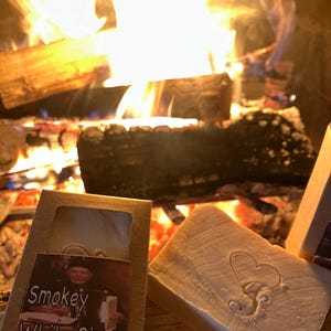 Puede incluir: Dos barras de jab&oacute;n hechas a mano, una con un dise&ntilde;o de coraz&oacute;n y la otra con una etiqueta que dice "Smokey White Birch" y "Woodlife Creations". Las barras de jab&oacute;n est&aacute;n frente a una chimenea con un fuego rugiente.