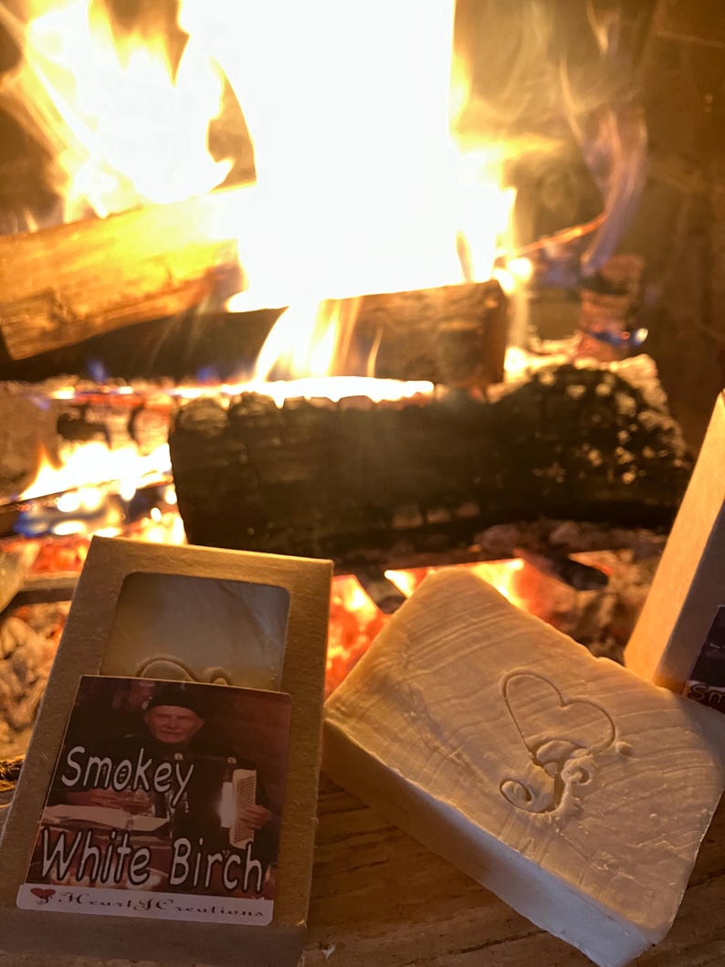 Puede incluir: Dos barras de jab&oacute;n hechas a mano, una con un dise&ntilde;o de coraz&oacute;n y la otra con una etiqueta que dice "Smokey White Birch" y "Woodlife Creations". Las barras de jab&oacute;n est&aacute;n frente a una chimenea con un fuego rugiente.