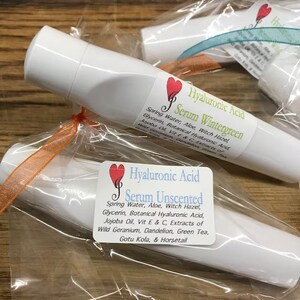 May include: Two white tubes of facial serum with labels. The label on the top tube reads "Hyaluronic Acid Serum Wintergreen" and lists ingredients. The label on the bottom tube reads "Hyaluronic Acid Serum Unscented" and lists ingredients.