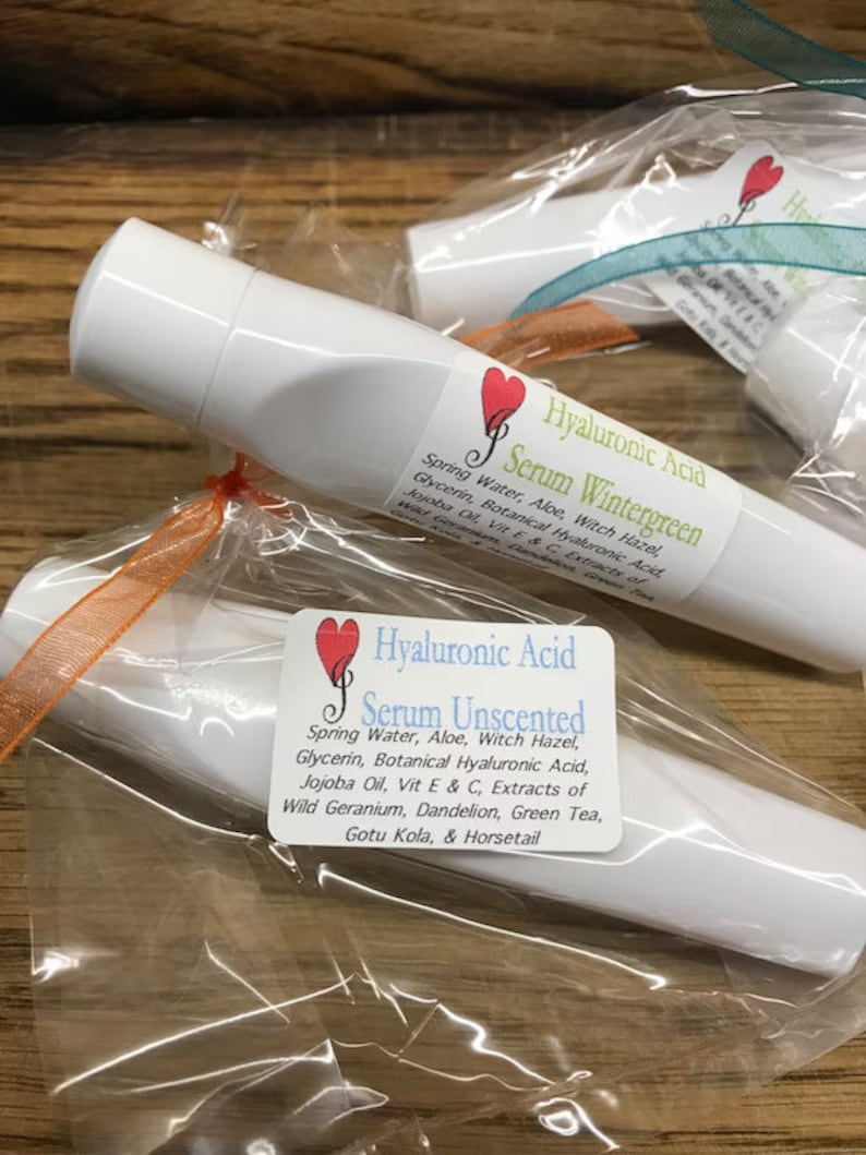 May include: Two white tubes of facial serum with labels. The label on the top tube reads "Hyaluronic Acid Serum Wintergreen" and lists ingredients. The label on the bottom tube reads "Hyaluronic Acid Serum Unscented" and lists ingredients.