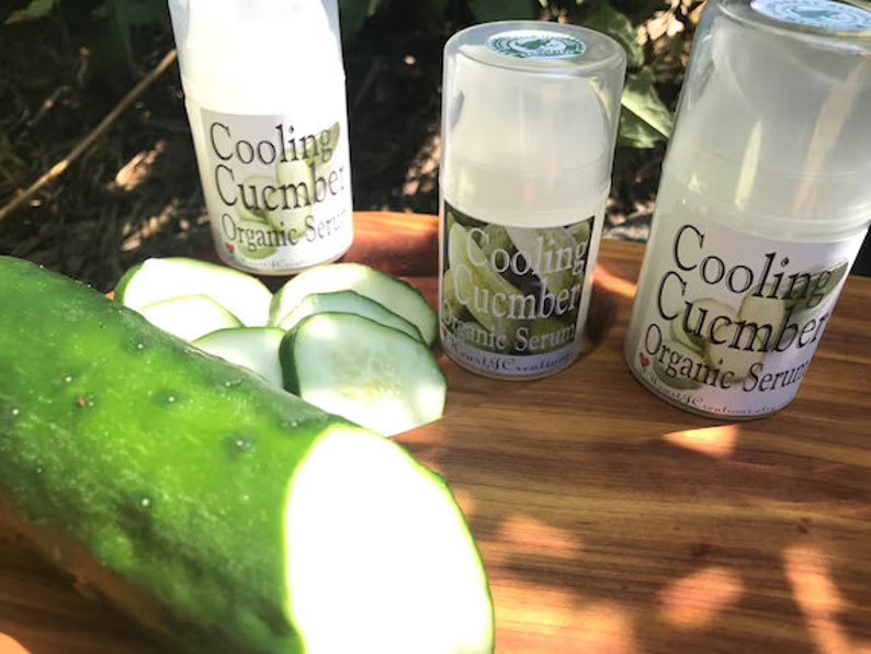 May include: Three small bottles of cooling cucumber organic serum on a wooden surface with a whole cucumber and sliced cucumbers.