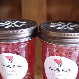 May include: Two glass jars filled with a red jelly-like substance. The jars have white lids with a decorative design and a white label with red hearts and the text "Smelly Jelly! Cherry Almond Fragrance Oils".