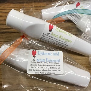 May include: Two white tubes of serum with labels that say "Hyaluronic Acid Serum Wintergreen" and "Hyaluronic Acid Serum Unscented". The labels have a red heart with a musical note in it. The tubes are wrapped in clear plastic with orange and teal ribbon.